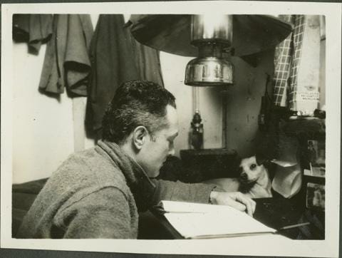 a man reading at a desk in a room with a light fixture above his head and a small dog laying on the desk in front of him. a man reading at a desk in a room with a light fixture above his head and a small dog laying on the desk in front of him.