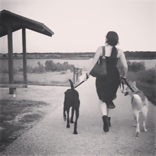 Old, black and white image of Lyric in their mid-20s. They are walking two large dogs (Dusty and Rockey) while wearing heels, a black dress, and a black purse over their arm. 