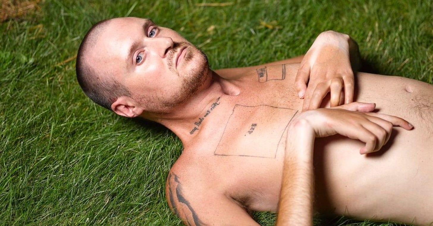 A white disabled man with tattoos is lying on grass, looking directly at the camera.