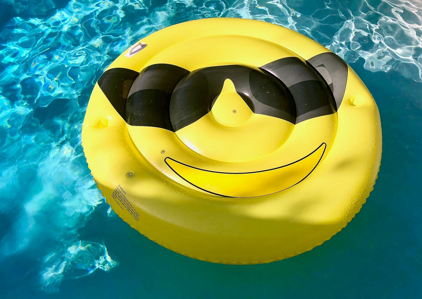 Piscina con sfondo azzurro e in superficie un materassino tondo di color giallo a forma di emoji sorridente con occhiali da sole.