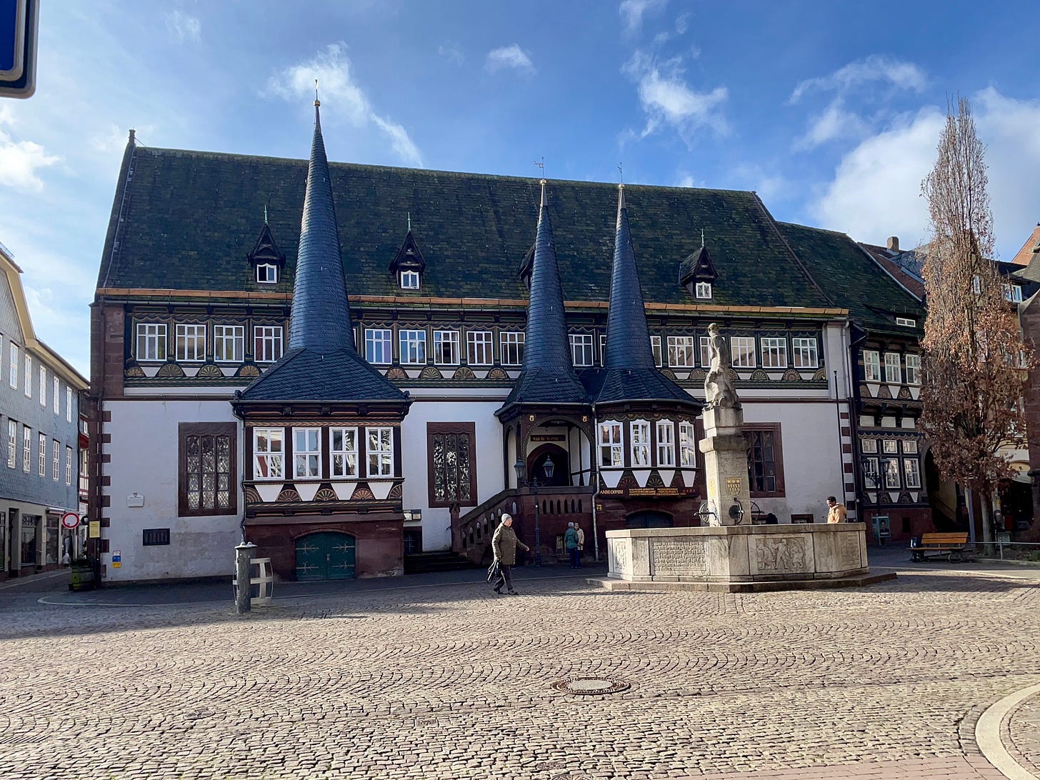 A historic half-timbered building with three distinctive pointed towers stands under a bright blue sky. The building, Rathaus Einbeck, features intricate woodwork, numerous windows, and a grand staircase leading to the entrance. In the foreground, a stone fountain with a statue adds to the charm of the cobblestone square, where a few people are walking. A historic half-timbered building with three distinctive pointed towers stands under a bright blue sky. The building, Rathaus Einbeck, features intricate woodwork, numerous windows, and a grand staircase leading to the entrance. In the foreground, a stone fountain with a statue adds to the charm of the cobblestone square, where a few people are walking.