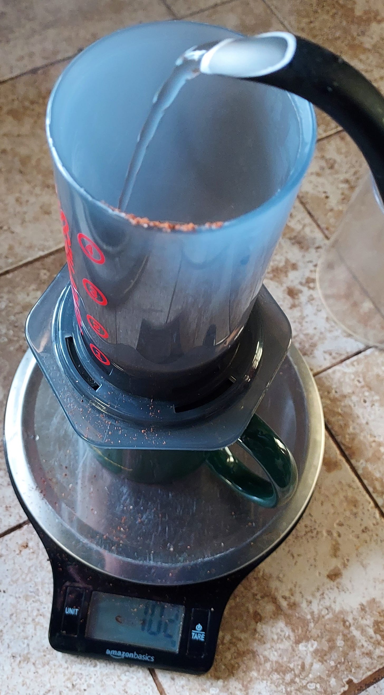 A top view of water being poured from a kettle spout into an Aeropress (grey plastic cylinder) coffee brewer that sits on a green coffee mug on top of a digital scale showing the weight in grams. The scale is black with a silver platform.