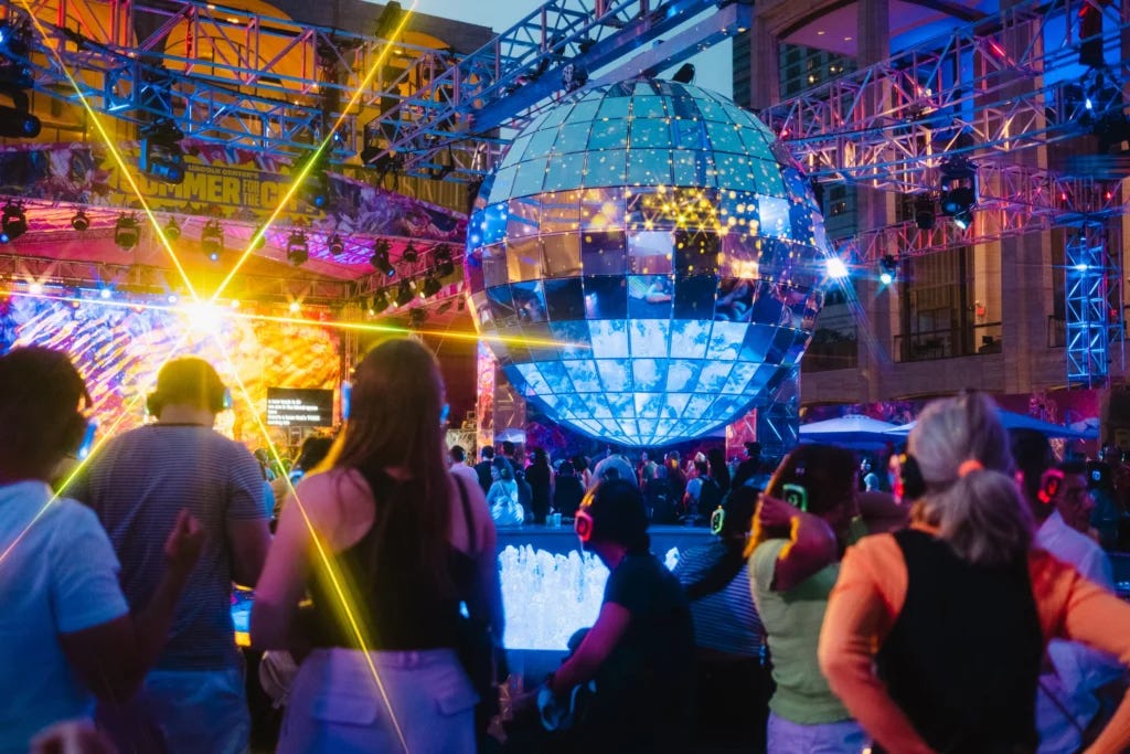 A photo from deep in a crowd of people wearing silent disco headphones at an outdoor party. They are looking up toward a stage in the distance. There is an enormous 10 foot tall disco ball hovering over the iconic fountains at Lincoln Center.