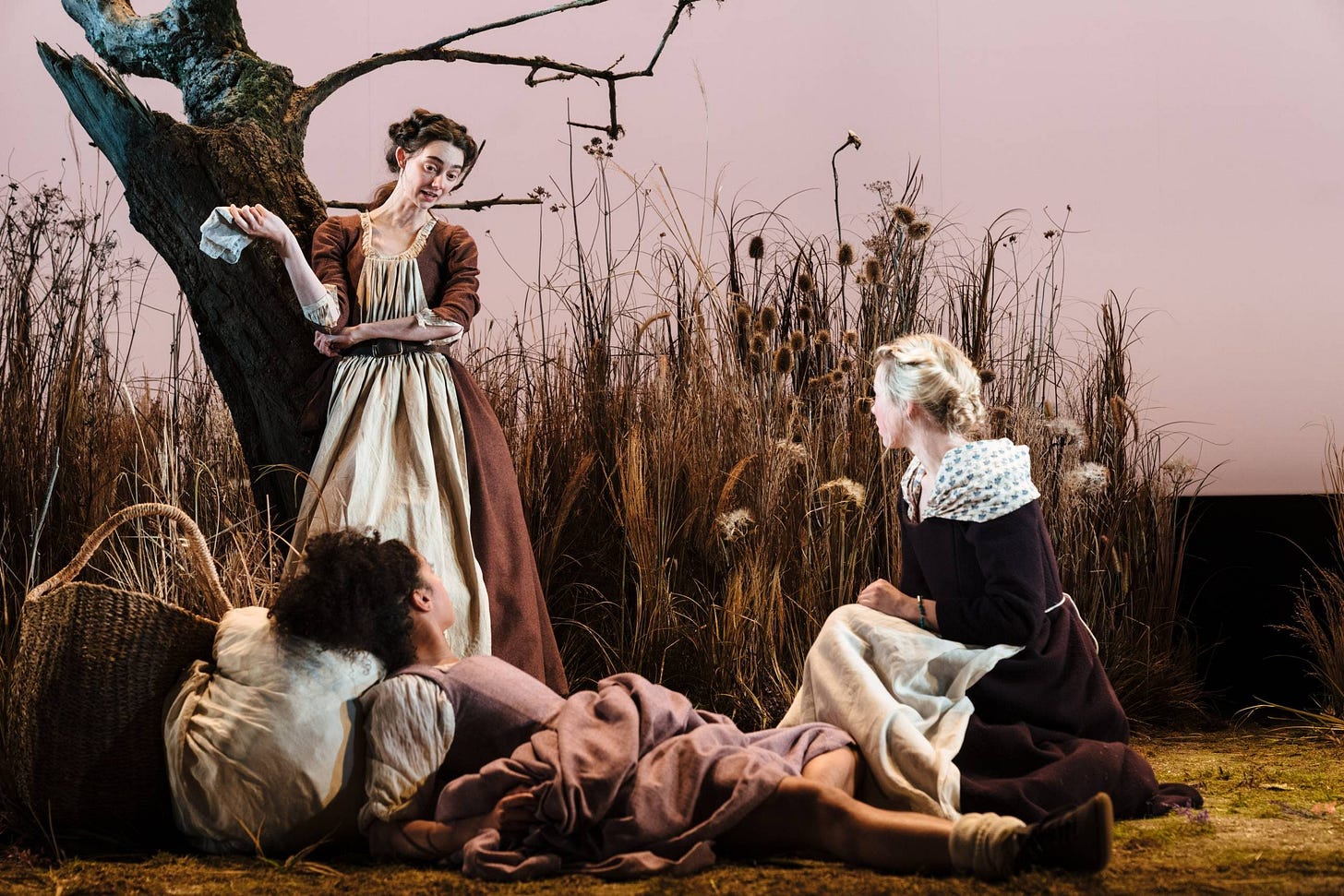 Liv Hill, Siena Kelly, and Tanya Reynolds are together in a field. Liv Hill wears a burgundy dress and white overskirt and crouches near Siena, who leans back on the ground against a laundry bundle, wearing a pink dress. Tanya talks to them while leaning against a tree. She wears a brown dress.