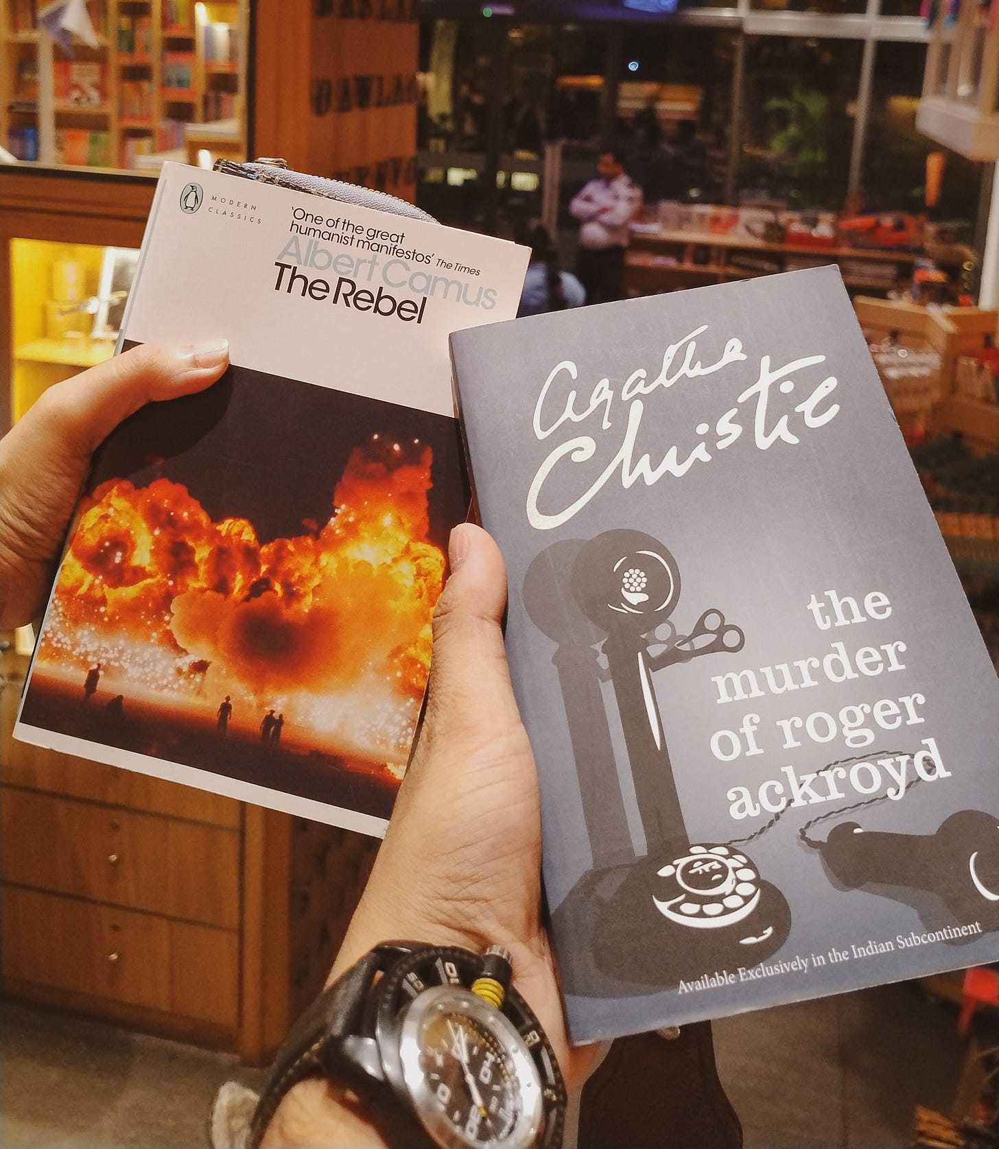 Two hands, holding The Rebel by Albert Camus and The Murder Of Roger Ackroyd by Agatha Christie inside a bookstore 