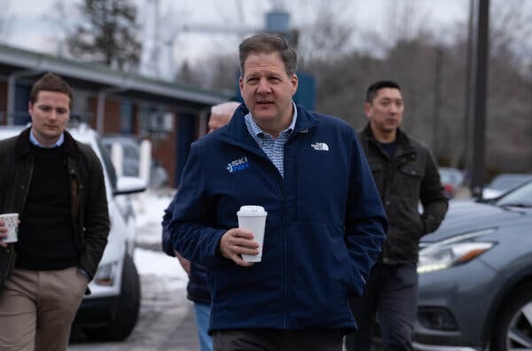 Governor Chris Sununu of New Hampshire walks through a parking lot while holding a to-go coffee cup, with several men walking behind him.