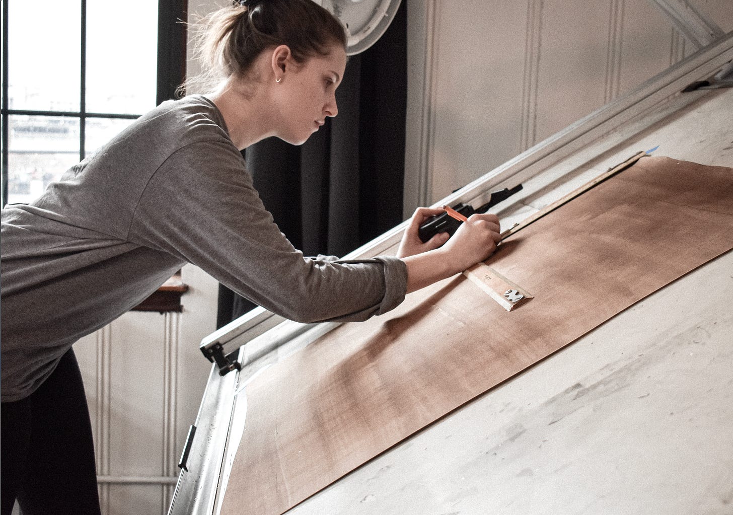 A photo of the artist measuring on canvas, leaning on her drafting table in front of her. She is wearing a grey shirt and has a bun in her hair, a window is behind her. 