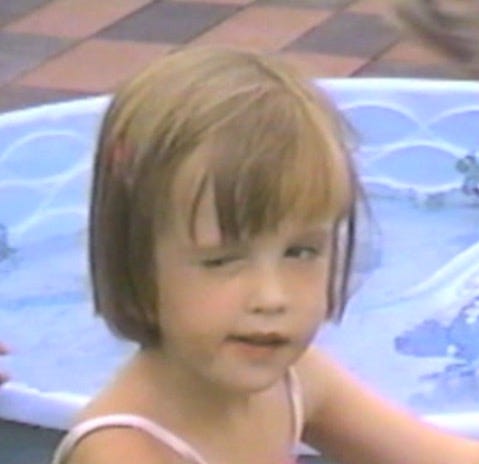 Me as a kid, sitting in a front of a plastic kiddie swimming pool and trying to wink. I'm white and have short hair with bangs. One eye is squeezed tight, and the other one is open but glancing toward the closed eye.