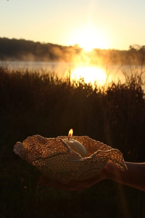 Woman holding a round lit candle inside a beautiful lace/crocheted bowl casting a warm glow in front of the reflection of the rising sun on a lake. Photo taken by Aleesha Boyd for Melanie Williams de Amaya.
