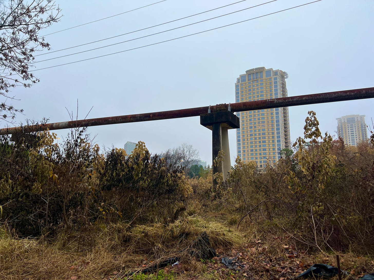 Color photo of Houston landscape: green-brown foliage in foreground, at the base of an elevated pipeline crossing the frame, power lines overhead, and two high rises in the background