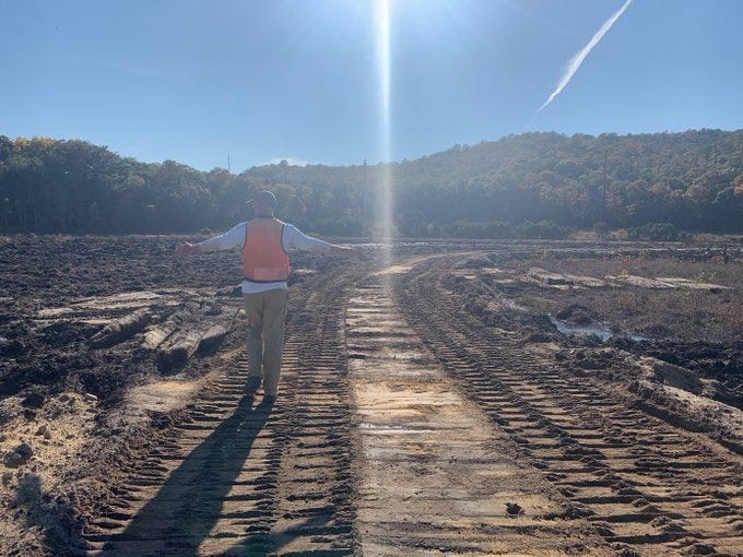 A muddy stretch of bare land with tracks from earthmoving equipment. A state worker in an orange safety vest stands in the seeming desolation with his arms stretched