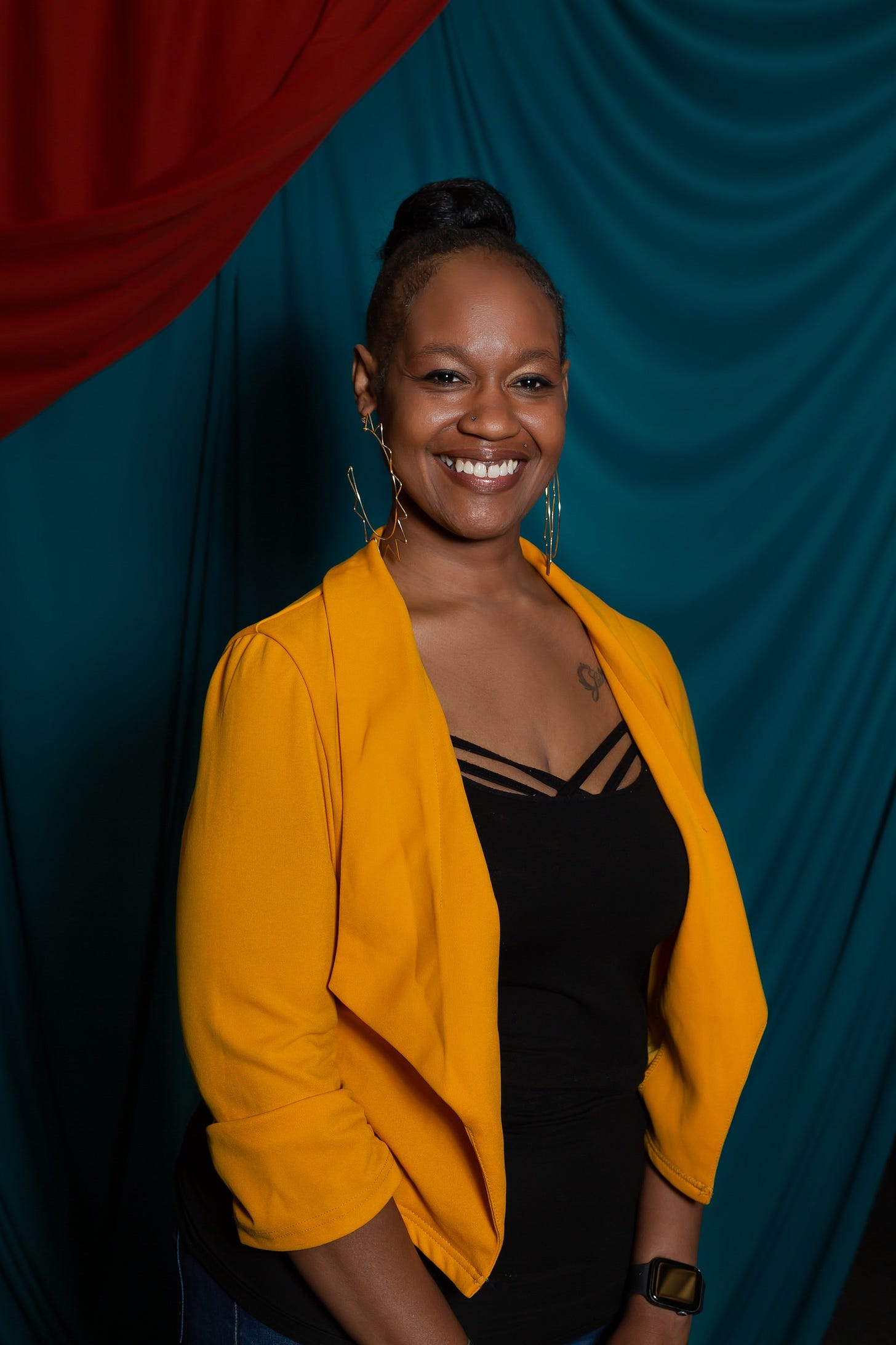 Portrait of a Black skinned woman, she is smiling at the camera. She has her hair up in a high bun. She is wearing large gold hoops and a large golden yellow blazer with a black shirt.