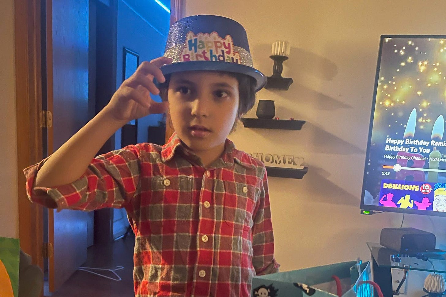 Wadee wears a Happy Birthday hat and holds a gift bag. A TV behind him displays a Happy Birthday message. 