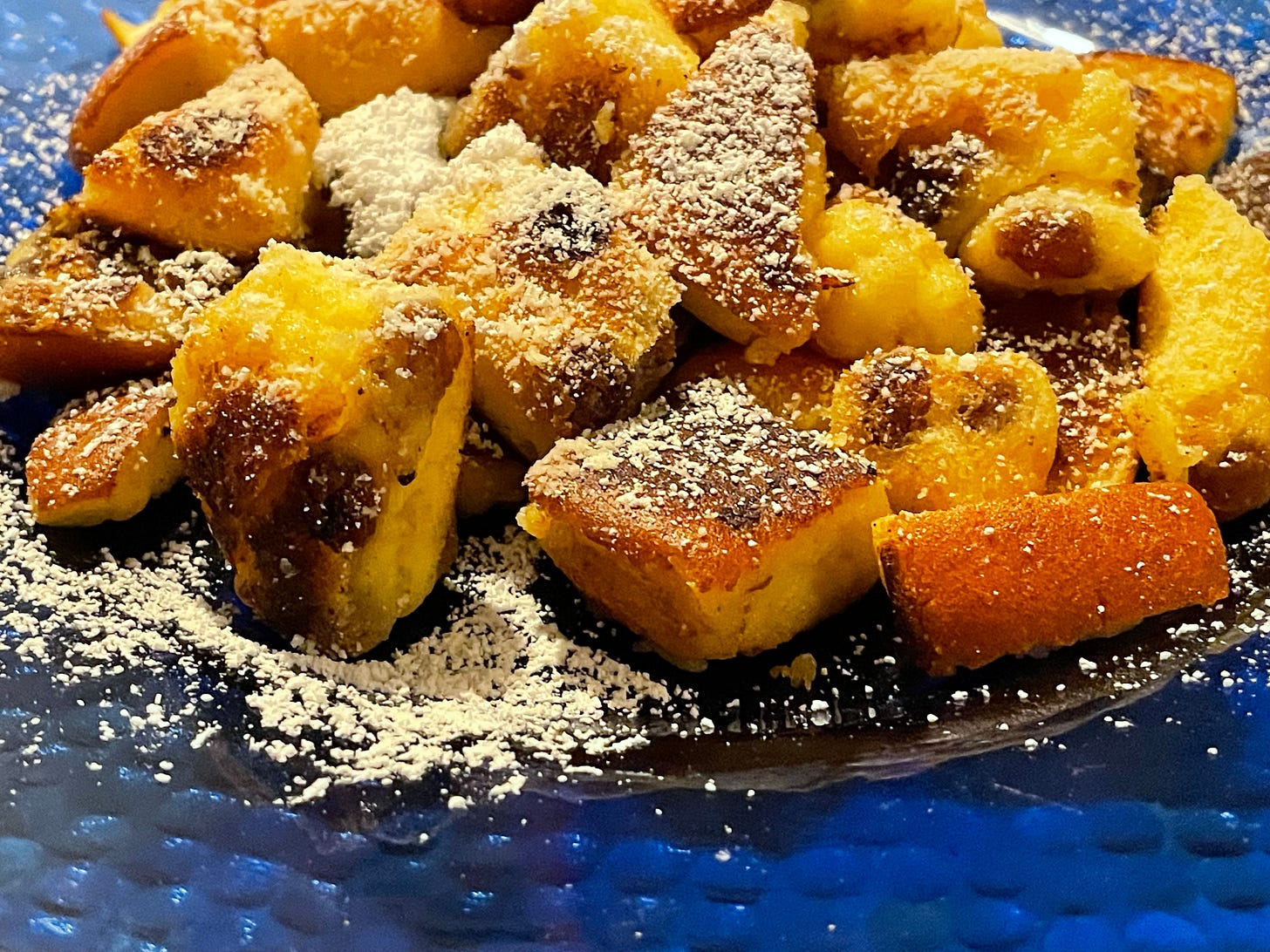 A serving of Kaiserschmarrn on a blue plate, dusted with powdered sugar. The golden-brown, fluffy pieces of torn pancake are lightly caramelized and scattered with raisins, giving a warm, inviting appearance. A serving of Kaiserschmarrn on a blue plate, dusted with powdered sugar. The golden-brown, fluffy pieces of torn pancake are lightly caramelized and scattered with raisins, giving a warm, inviting appearance.