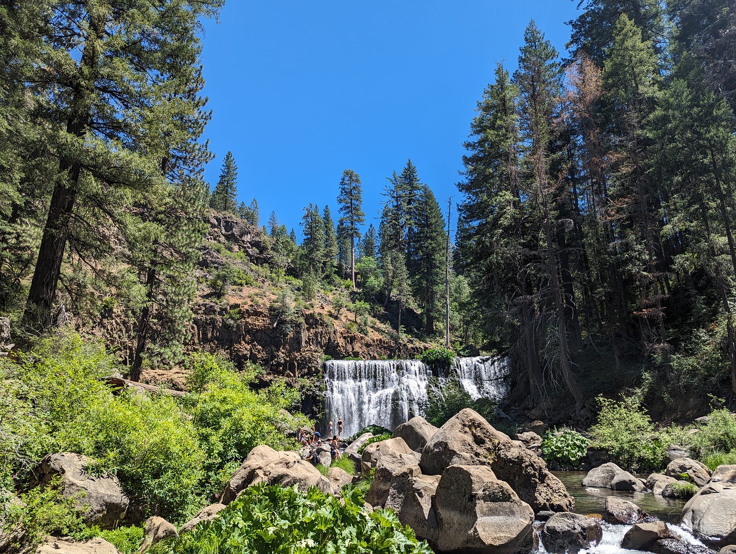 Amid a forest of green pine is a boulder-strewn river that wends its way from a cascading waterfall in the near distance. People play and bathe in the cold pools that have formed in the river, enjoying the chill water in the hot, blue-skied day. 