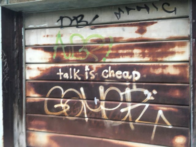 An attractively rusted, otherwise white-painted garage door in a Toronto alley, with various graffiti including the words, in small writing, Talk is cheap