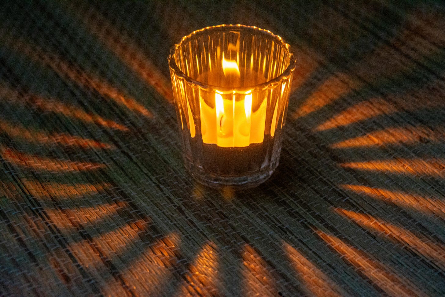 A imagem mostra uma vela acesa dentro de um copo de vidro. A chama brilha intensamente, iluminando o ambiente. O copo de vidro possui ranhuras verticais, que criam um interessante padrão de luz e sombra ao redor, projetado sobre o chão. Este chão parece ser feito de um material trançado, como uma esteira de palha A imagem mostra uma vela acesa dentro de um copo de vidro. A chama brilha intensamente, iluminando o ambiente. O copo de vidro possui ranhuras verticais, que criam um interessante padrão de luz e sombra ao redor, projetado sobre o chão. Este chão parece ser feito de um material trançado, como uma esteira de palha