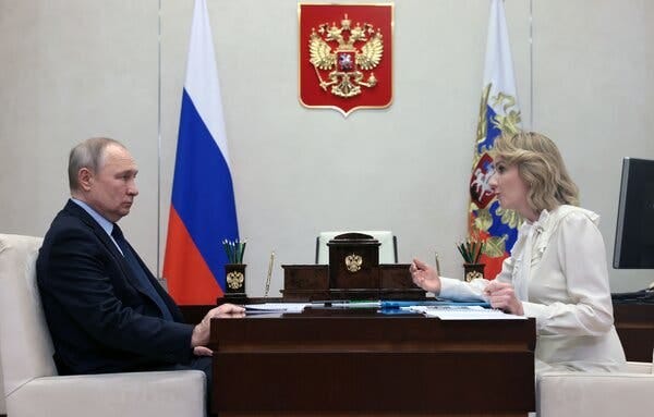 Russia's leader dressed in a dark suit and seated across the table from a government official dressed in white. Flags are displayed in the background.
