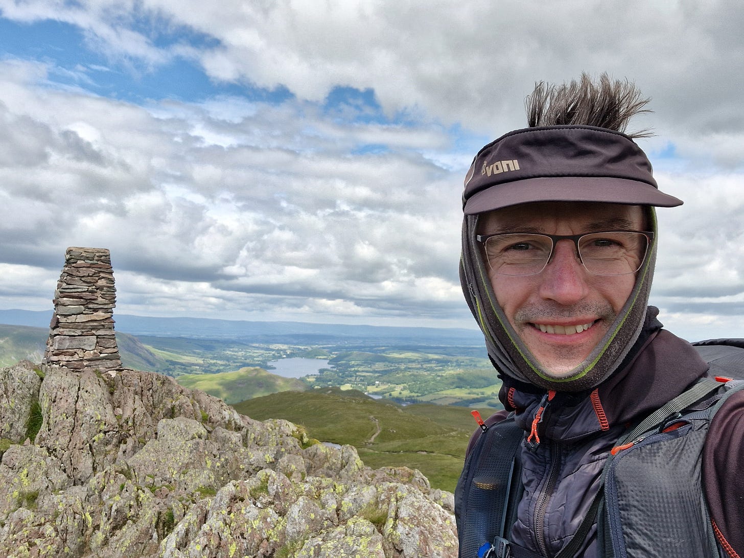 Summit of Place Fell with stone pillar and lake (Ullswater) behind with man in foreground.