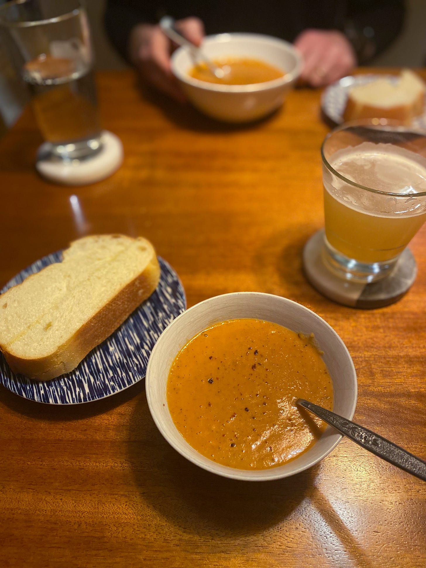 A small white bowl of the tomato red lentil soup described above, with a spoon in it. On a blue and white plate to the left is a slice of garlic bread, and to the right is a glass of IPA. The same meal sits in front of Jeff on the opposite side of the table. His hand curls around the edge of his bowl.
