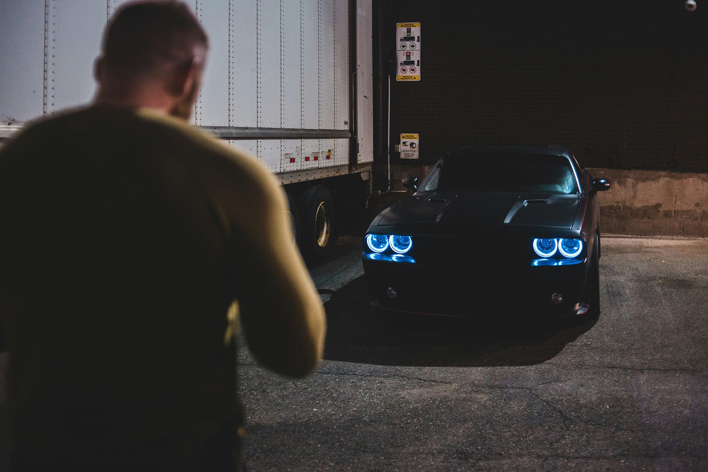 A man stands in front of a black car with a semi-trailer beside the car, at night.