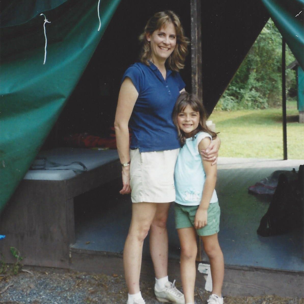 Young mom stands with her 7-year-old daughter by a tent on a mother-daughter camping trip
