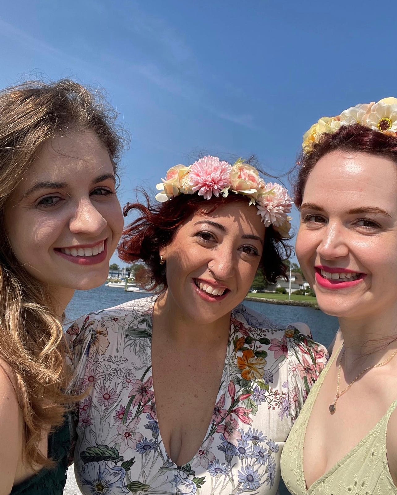 Three white women with flower crowns on are standing in front of an ocean, smiling. 