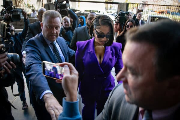 Megan Thee Stallion, in a bright purple pantsuit and sunglasses, being escorted into a courthouse.