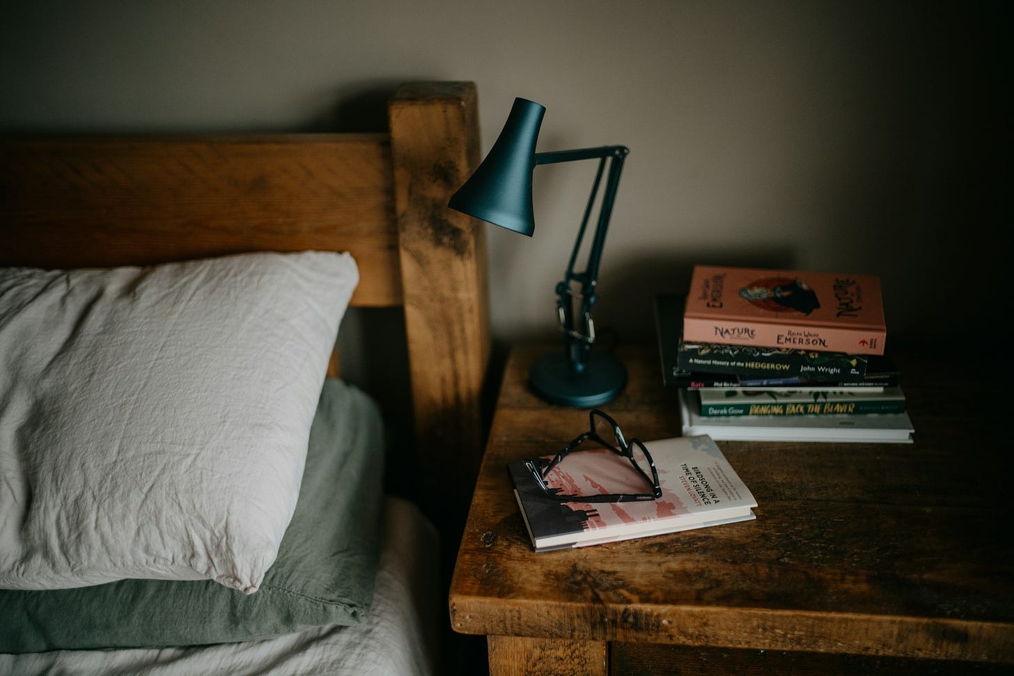 The top part of a bed with pillows and a headboard, next to a bedside table with a bedside lamp, a stack of books, and reading glasses on it