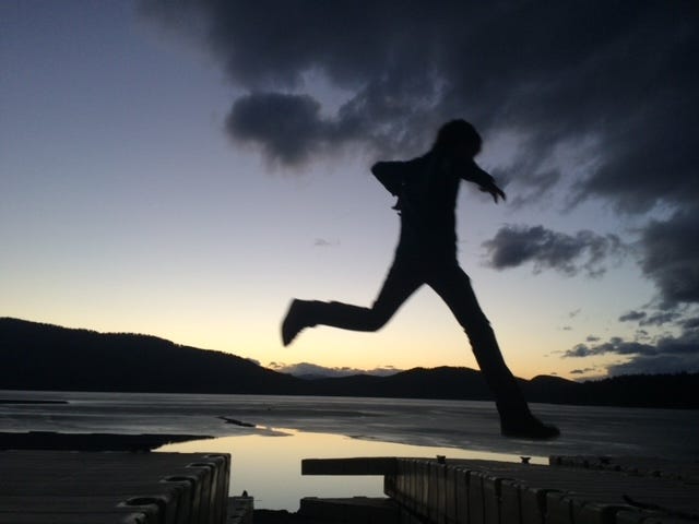 The silhouette of a boy jumping across a dock with the lake and mountains in the background.