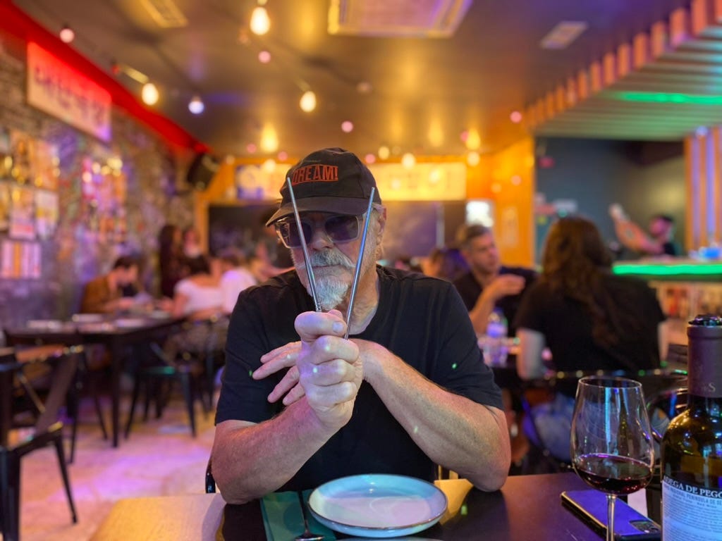 Man with ace moustache and beard wearing a cap and holding chopsticks in a Korean restaurant