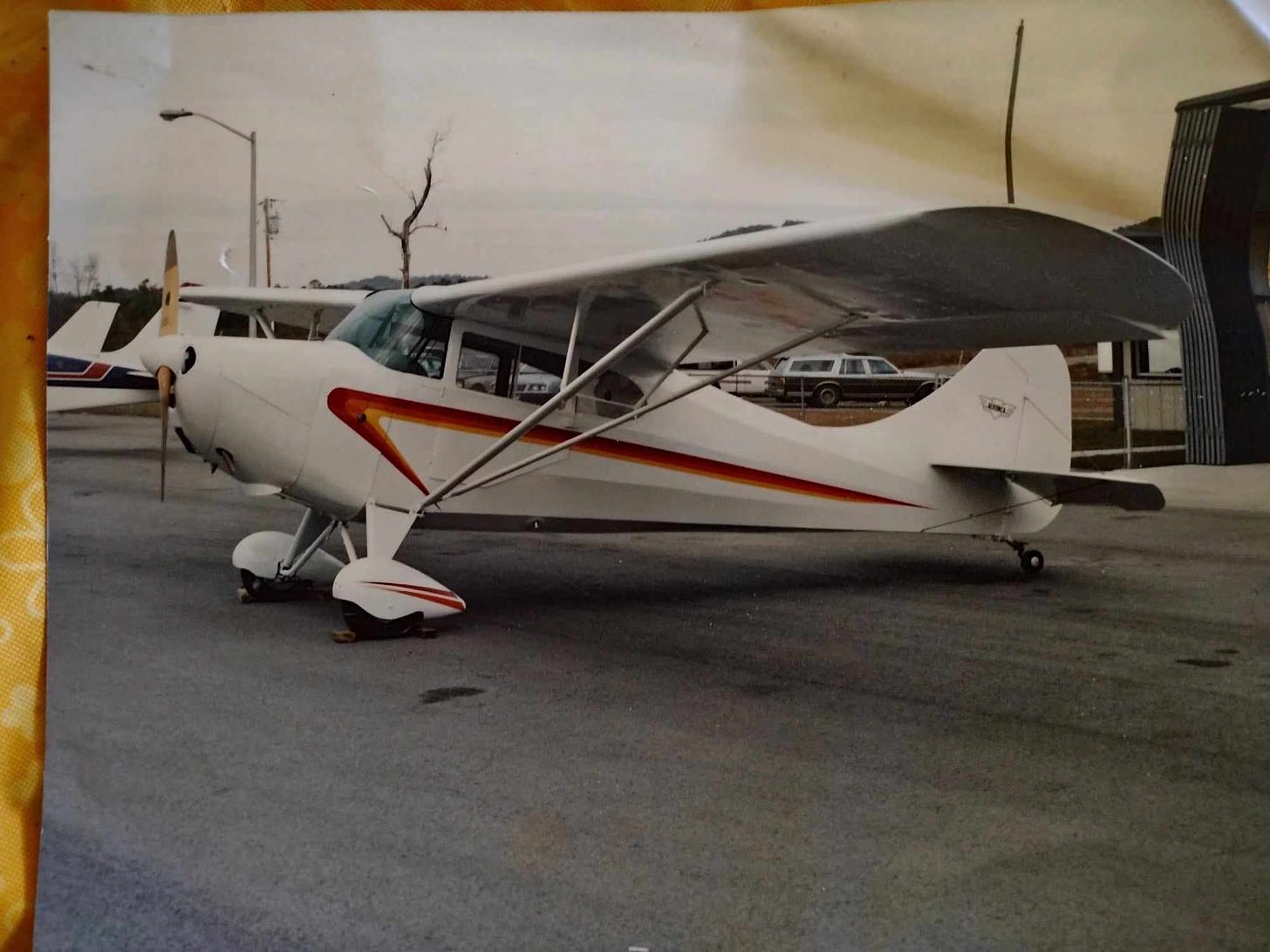 A small plane, with a propeller, and a red, orange, and yellow stripe down its side, sitting in what looks like a parking lot. The image is of an old photo, and there's a station wagon in the background, and what appears to be a dead tree, and a light post. You can also see the tail fin of another plane. 