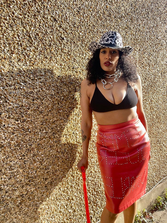 An image of a femme person of colour with long curly hair. They wear a black bra top and a red bedazzled skirt and a cowprint stetson hat. They lean on their red walking stick.