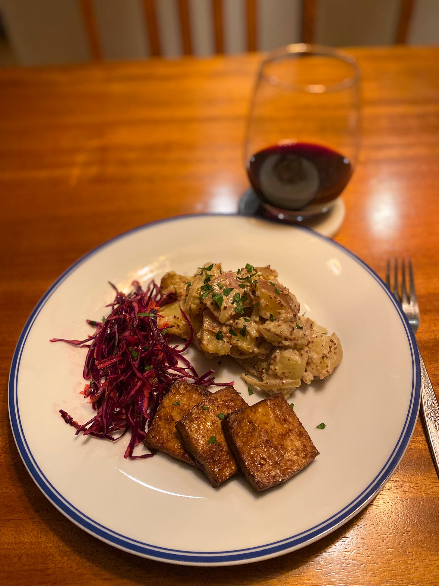A dinner plate with three large squares of dark brown marinated tofu arranged next to a grainy mustard potato salad in the upper right, and a red cabbage and carrot slaw on the left. On a coaster behind and to the right of the plate is a stemless glass of red wine.