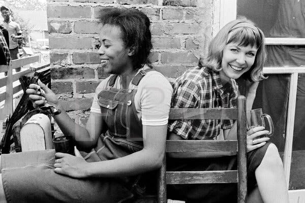 Two young women sitting back to back and smiling. The woman on the left is Black and wears overalls; the woman on the right is white, with blond hair, and is wearing a plaid shirt and a dark skirt.