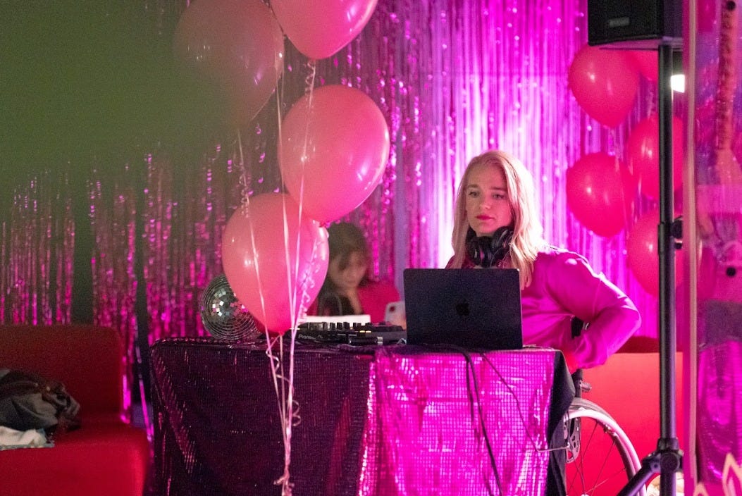 Surrounded by pink balloons and pink curtains, I am DJing the Barbie opening party. I am also dressed entirely in pink, including pink hair and a pink lip. 
