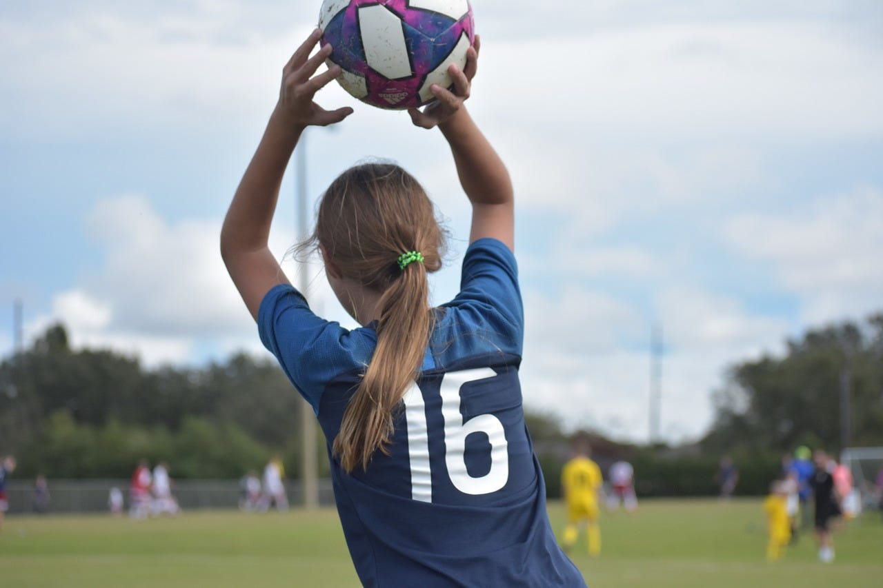 Photo of a girl throwing a soccer ball. It is shot from the back and other players are out of focus.