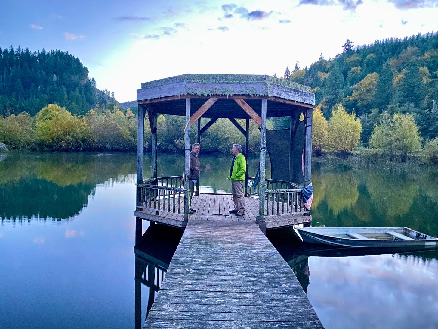 A wooden dock leads to a covered gazebo on a pond at sunset, with two men talking to each other in the gazebo.