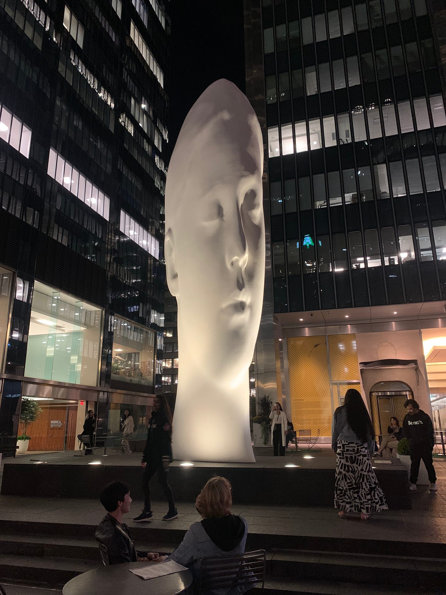A sculpture of an elongated white head stretching up into the night sky.