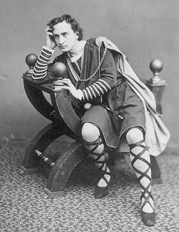 An old black and white photo of the actor Edwin Book as Hamlet. He sits looking sulky and resentful and brooding on a stool. He wears a tunic and long laces up his hose to the knee.
