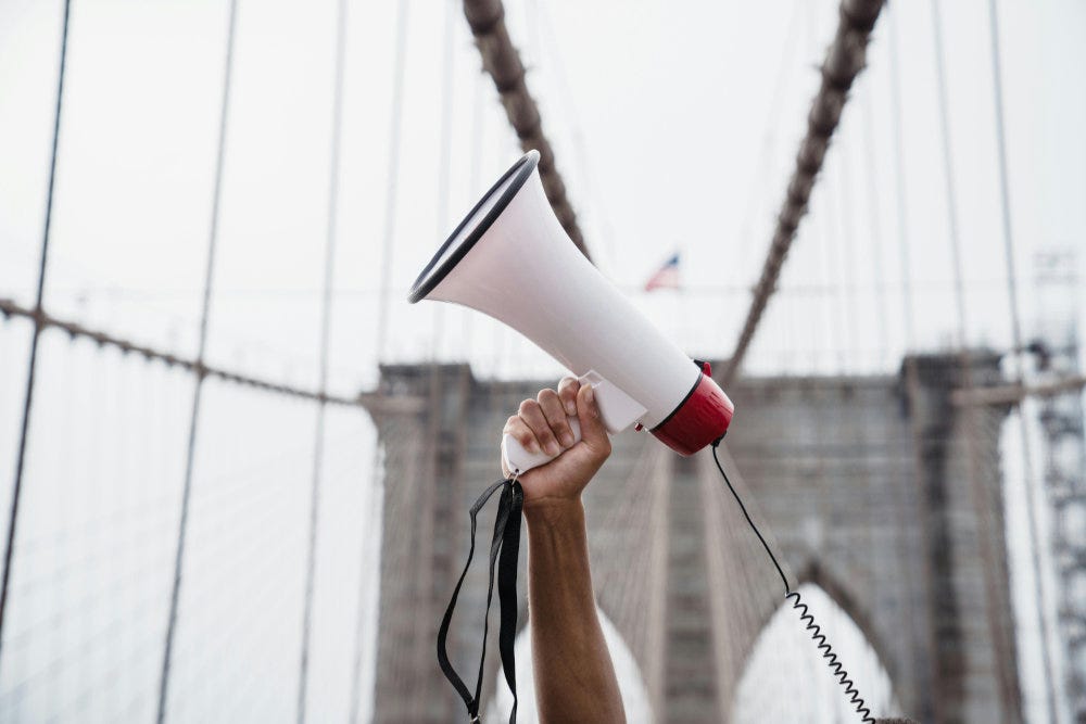 A hand holds a megaphone in the air in front of the cables and stone tower of a suspension bridge.