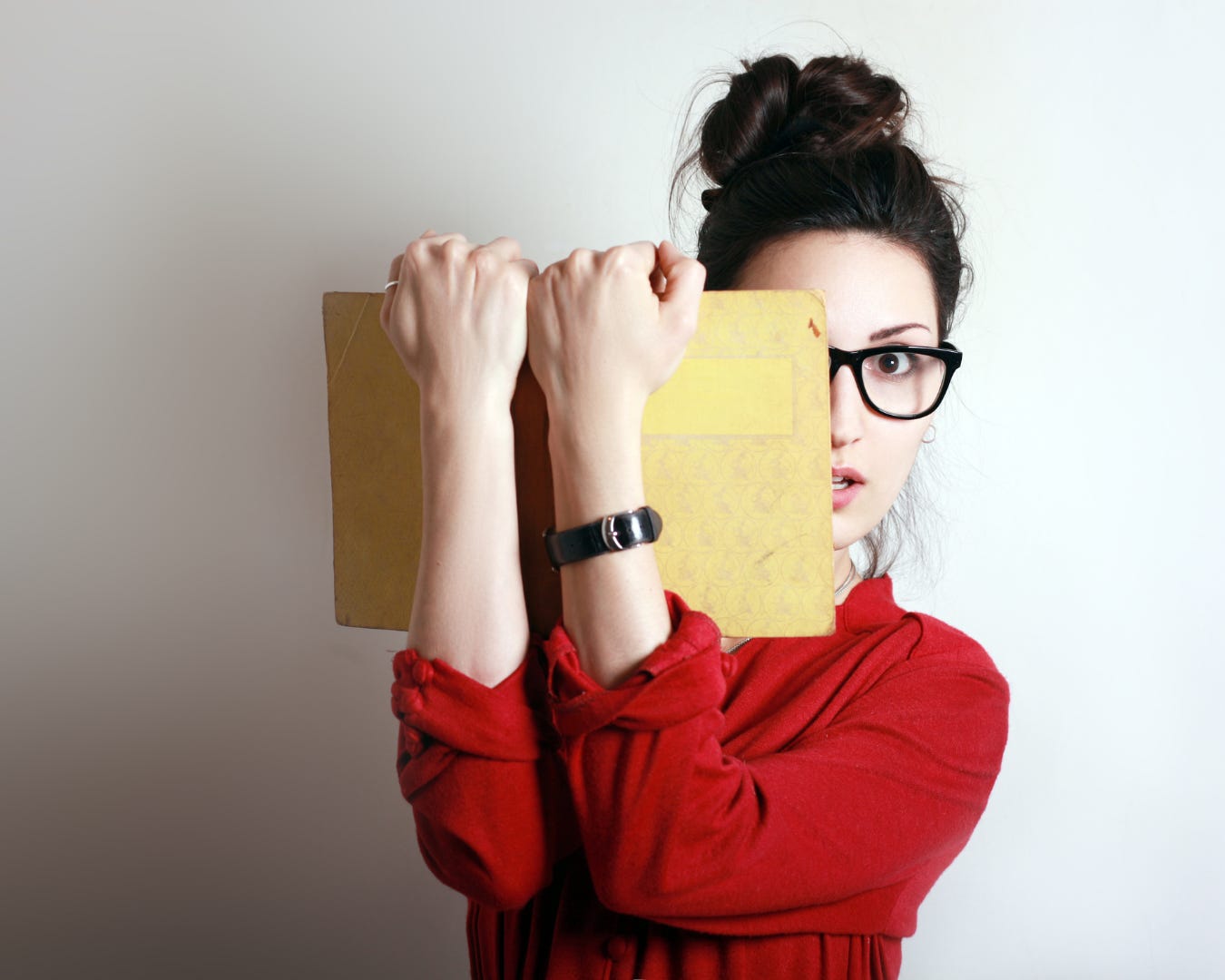 A woman wearing glasses partially hides her face behind the book she is holding in front of her.