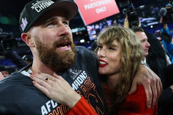 Travis Kelce, in a black hat and black shirt, has his arm around Taylor Swift who is wearing a red top and two rings.