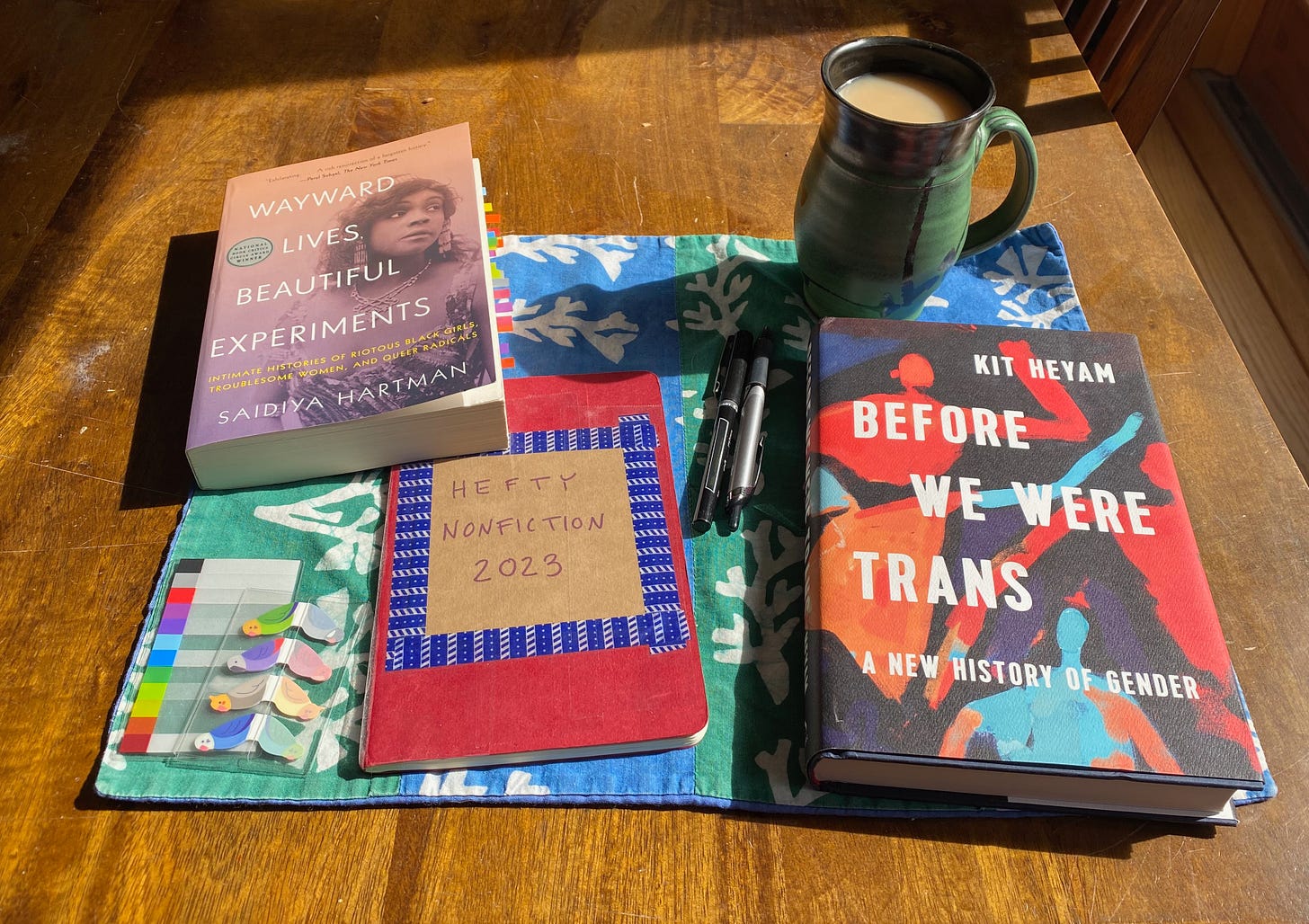 Before We Were Trans & Wayward Lives, Beautiful Experiments sit on a colorful placemat next to a red notebook titled 'Hefty Nonfiction 2023', a few pens, a pack of colorful tabs, and and a mug of tea.