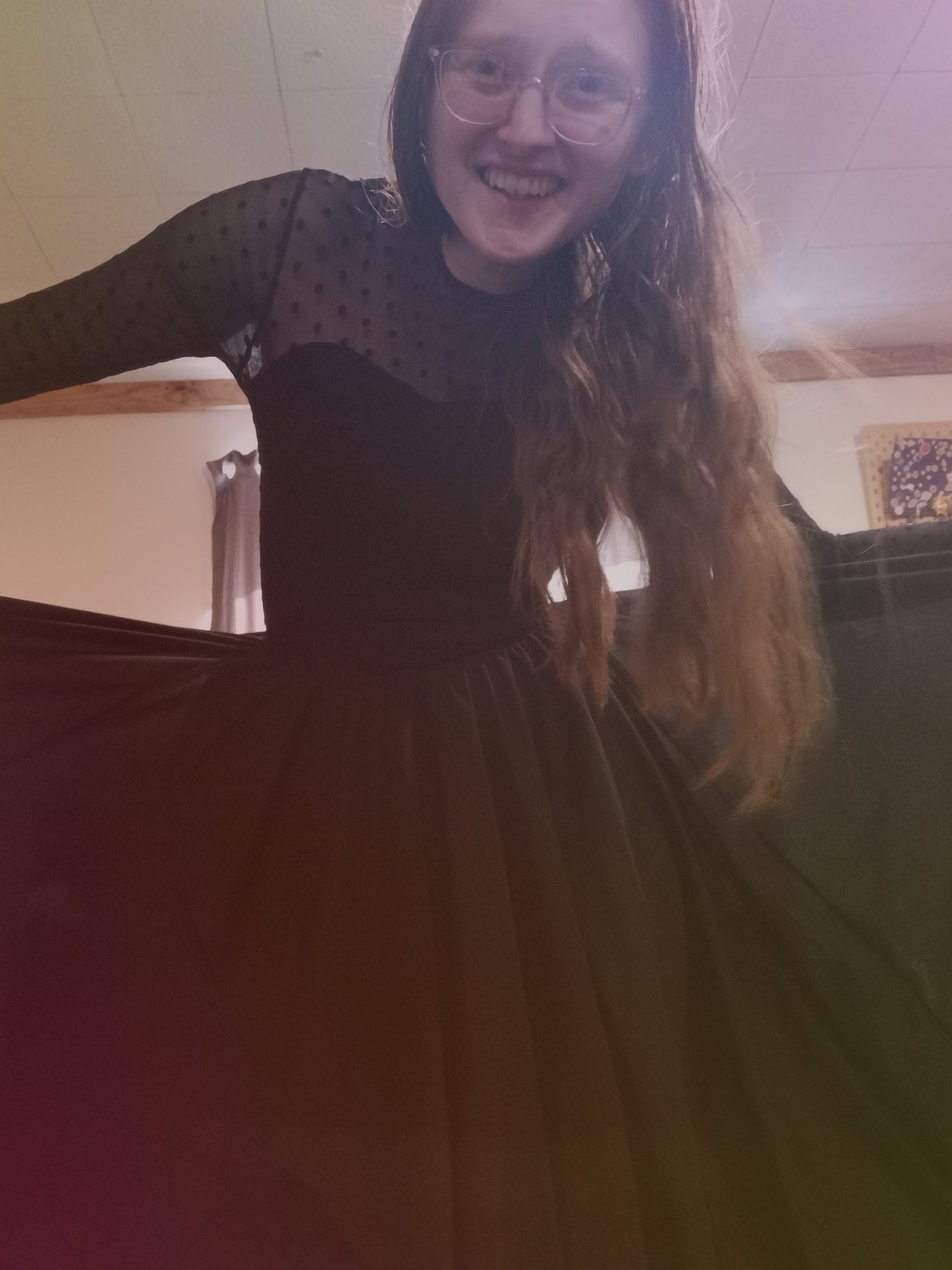 Kandi Zeller (She/Her), a white woman with long reddish brown hair, smiles at the camera while wearing a flowy black dress with a sweetheart neckline and a bodice covered in mesh with polka dots. 