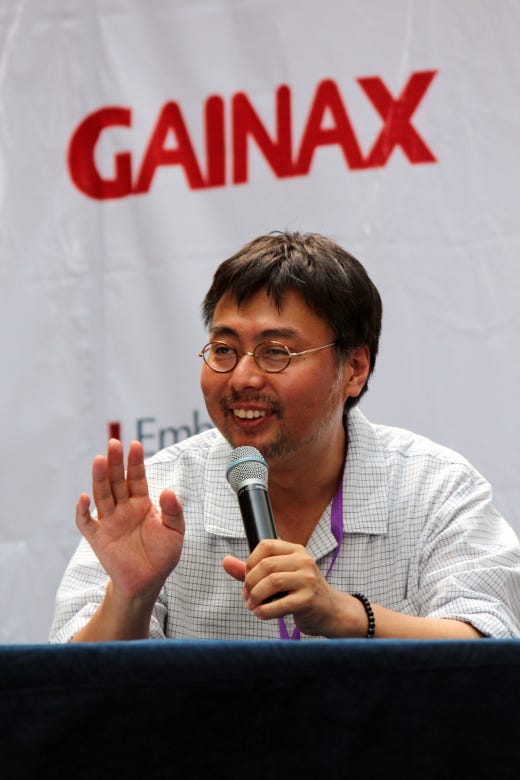 A man with round eyeglasses sits behind a table, holding a microphone and waving to people offscreen. Above him in the background is the word Gainax printed on a banner.