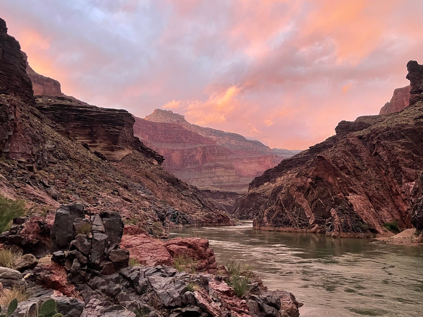 The Grand Canyon at sunset. Pink and orange clouds streak the sky above pink and gray rock and a green river.