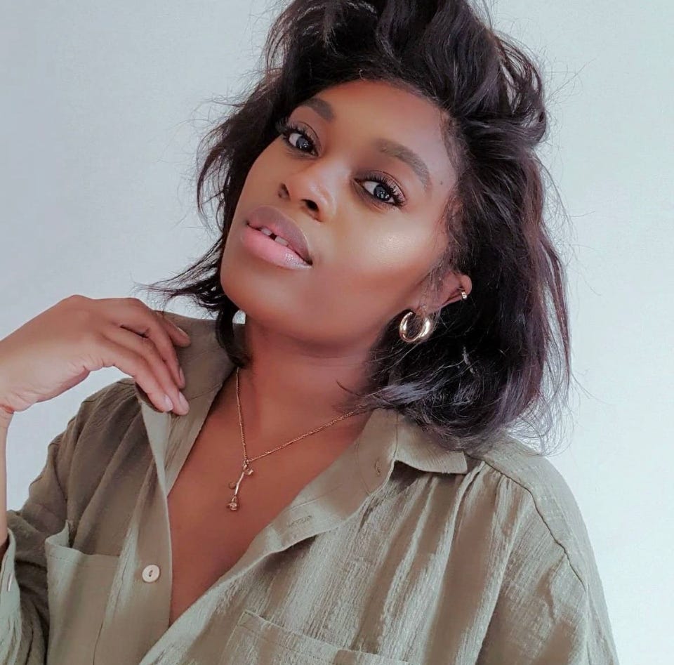 A black woman wirth short black hair wearing a khaki green collared button up shirt. She is also wearing gold hoop earrings & a gold necklace with a delicate rose as a pendant.