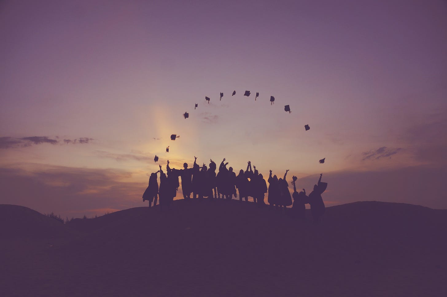 Silhouette of graduates throwing caps in the air. The caps, which are black, form an arc set against the backdrop of a purplish-yellow sky.