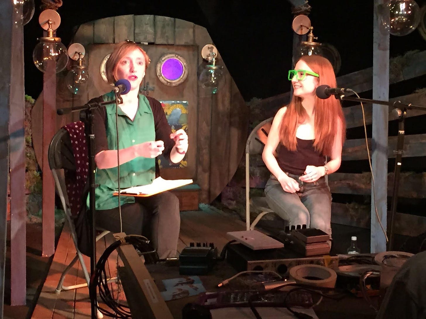 Two white women (one with short fair hair and one with long ginger hair)sit on chairs with microphones with an elaborate stage set behind them. 